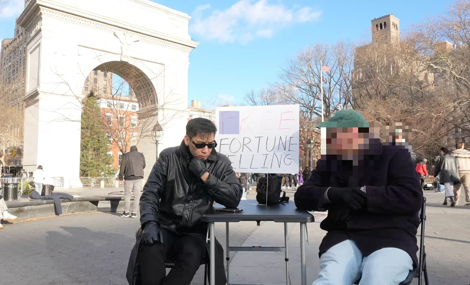 Ricky Li giving a free in person Chinese astrology reading at a table in Washington Square Park, New York, with the marble arch behind him.